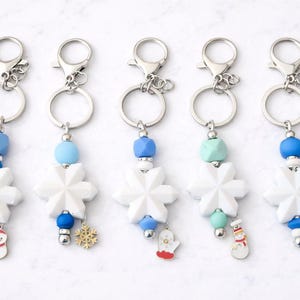 May include: Five snowflake keychains with silver rings and clasps. Each keychain has a white snowflake charm, colorful beads, and a winter-themed charm, including a snowman, snowflake, and mitten.