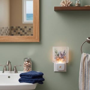 May include: A decorative night light featuring a seashell design, plugged into a wall outlet. The night light has a square, frosted glass cover with a coastal theme. The bathroom setting includes a sink, towels, and a jar of seashells.