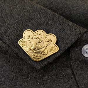Puede incluir: Un pin de metal dorado con un diseño intrincado, que presenta un emblema central y texto circundante. El pin está sujeto a una tela gris oscuro, probablemente una camisa o chaqueta. El diseño del pin incluye una imagen estilizada y símbolos.