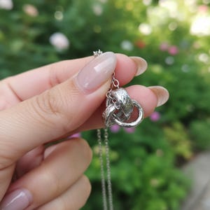 Puede incluir: Un collar plateado con un colgante de tres anillos entrelazados. Los anillos tienen un diseño ornamentado y texturizado. El collar se muestra sobre un fondo verde borroso, posiblemente al aire libre.