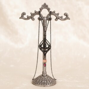 May include: A silver-colored jewelry display stand with ornate scrollwork. A black necklace with a pendant and a chain hangs from the top. A pink ring with a red stone is also displayed. The stand has a decorative base.