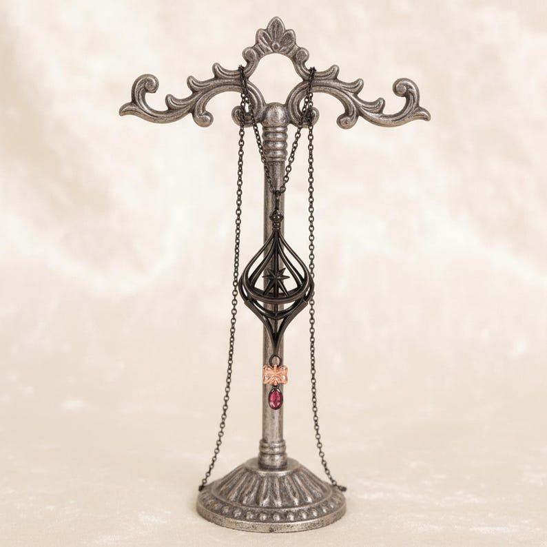 May include: A silver-colored jewelry display stand with ornate scrollwork. A black necklace with a pendant and a chain hangs from the top. A pink ring with a red stone is also displayed. The stand has a decorative base.