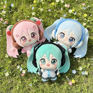 May include: Three plush dolls featuring large eyes and stylized hair. One doll has pink hair with cherry accents, one has light blue hair with snowflake details, and one has teal hair with black headphones, all wearing outfits with distinct color schemes.