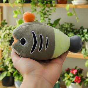 May include: A plush fish toy in shades of green, gray, and orange. The toy has a rounded body with black and white details, including a large eye and three curved lines. An orange ball sits atop the fish's head. The fish is held in a hand.