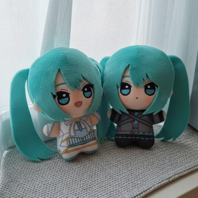 May include: Two plush dolls with teal hair and large blue eyes. One doll wears a white and blue outfit, the other a grey and black outfit. Both dolls have teal pigtails and are positioned side by side.