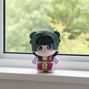 Pu&ograve; includere: Una bambola di peluche con capelli verdi, occhi blu e un vestito rosa e viola. La bambola &egrave; in piedi su una superficie bianca davanti a una finestra con uno sfondo verde sfocato. La bambola ha un aspetto carino e da cartone animato.