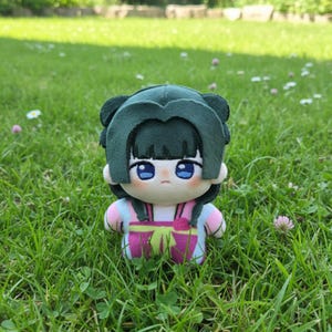 Pu&ograve; includere: Una bambola di peluche con una copertura per la testa verde a forma di capelli e un abito rosa e viola. La bambola ha grandi occhi blu ed &egrave; seduta in un'area erbosa. L'abito della bambola ha un dettaglio a fiocco.