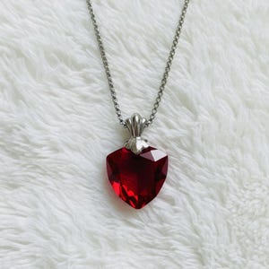 Può includere: Una collana color argento con un ciondolo a forma di cuore sfaccettato rosso intenso. Il ciondolo è sospeso a una delicata catena e ha un'ansa decorativa argentata. La collana è esposta su una superficie bianca strutturata.