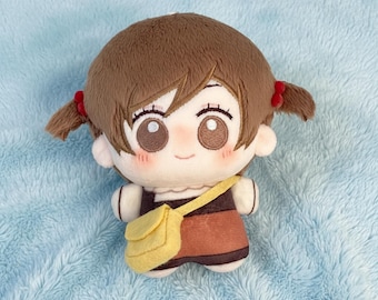 May include: A plush doll with brown pigtails, each tied with a red bow. The doll features large eyes, rosy cheeks, and a small yellow satchel. The doll is dressed in a brown and orange outfit.