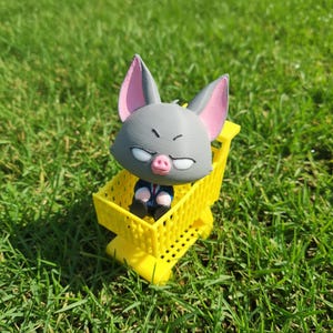 Puede incluir: Una caprichosa figura impresa en 3D de un murciélago gris con orejas rosas y una nariz rosa, vistiendo un traje, sentado en un carrito de compras amarillo brillante en miniatura. La figura está sobre un fondo de hierba verde.