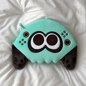 May include: A plush pillow shaped like a video game controller in mint green and dark brown. The center features two large white and black circular eyes. The surface includes embroidered directional buttons, a joystick, and X, Y, A, B letter buttons.