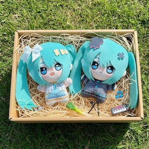 May include: Two plush dolls with turquoise hair and large eyes are presented in a wooden box filled with straw. One doll has a white bow, the other a cross-shaped hair accessory. Miniature musical instruments and a leek are also in the box.