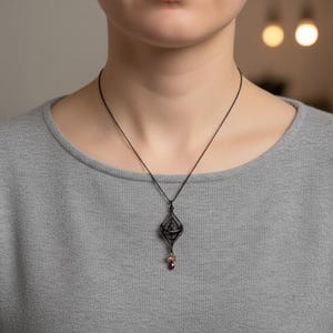 May include: A dark metal pendant necklace with a geometric design and a small, dangling red gemstone. The necklace is worn on a gray shirt. The pendant has an intricate, openwork design.