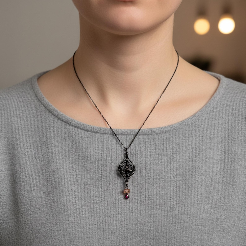 May include: A dark metal pendant necklace with a geometric design and a small, dangling red gemstone. The necklace is worn on a gray shirt. The pendant has an intricate, openwork design.