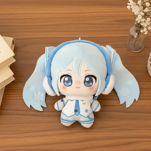 May include: A plush doll featuring light blue hair in long twin tails with white snowflake embroidery, wearing a white and blue outfit with a matching scarf and headphones. The character has large blue eyes, a small mouth, and rosy cheeks.