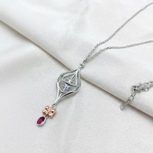 May include: A silver pendant necklace with a unique lantern-like design. The pendant features a cage-like structure with a star-shaped accent and a small, red gemstone. A small, rose-gold bow detail adds a touch of elegance. The necklace is displayed on a white, silky background.