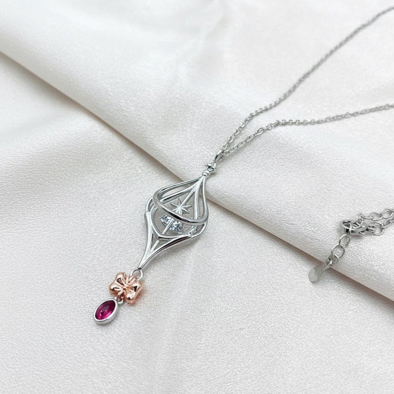 May include: A silver pendant necklace with a unique lantern-like design. The pendant features a cage-like structure with a star-shaped accent and a small, red gemstone. A small, rose-gold bow detail adds a touch of elegance. The necklace is displayed on a white, silky background.