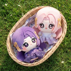 Puede incluir: Dos muñecas de peluche con ojos grandes y trajes detallados están sentadas en una cesta de mimbre sobre hierba. Una muñeca tiene el pelo morado y un vestido a juego, mientras que la otra tiene el pelo rosa y un vestido lila claro. Las muñecas tienen detalles dorados.