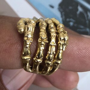 May include: A gold-toned ring featuring a multi-layered design that resembles a skeletal hand wrapping around the finger. The band is composed of several articulated segments, each detailed with small, raised skull motifs and bone-like structures.