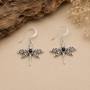 Può includere: Un paio di orecchini a forma di libellula in argento. Ogni orecchino presenta un design dettagliato a libellula con ali traforate e una pietra nera al centro. Gli orecchini hanno una chiusura a gancio.