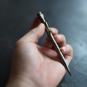Op de afbeelding: Een slank, metalen vulpotlood met een donkere, verweerde afwerking. Het schrijfinstrument heeft een taps toelopende punt, een cilindrisch lichaam en een kleine, geïntegreerde zakclip met een afgerond uiteinde bij de dop.