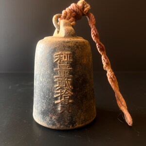 May include: A weathered, dark gray bell with embossed Japanese characters. A twisted, reddish-brown rope is attached to the top. The bell has a loop for hanging and shows signs of age and use.