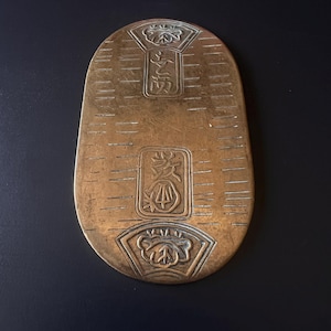 Può includere: Un oggetto metallico oblungo color bronzo con tre decorazioni floreali e caratteri giapponesi. La superficie presenta linee orizzontali. L'oggetto è su uno sfondo scuro.