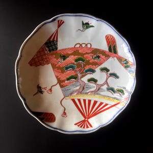 May include: A white ceramic plate with a scalloped edge, featuring a Japanese-style landscape design. The design includes a red fan, trees, and birds, with a blue border. The plate is approximately 8 inches in diameter.