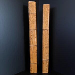 May include: Two tall, bamboo-style decorative columns. The columns are a light tan colour with darker brown markings, resembling natural bamboo. Each column is segmented into sections, adding to the bamboo aesthetic. They stand against a dark blue wall.