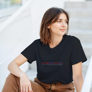 May include: Black t-shirt with red and pink text: "pd.merge(me, you, how='inner') I'm only complete when we are together." The shirt is a crew neck style. The person is wearing brown trousers.