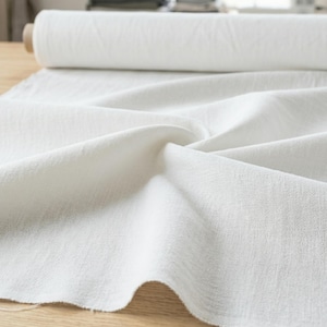 May include: A roll of white fabric sits on a wooden surface, with a portion of the fabric unrolled and draped. The fabric has a textured appearance and is likely linen or a similar material. The background includes a sewing machine and other fabric.
