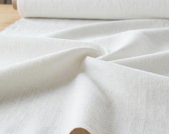 May include: A roll of white fabric sits on a wooden surface, with a portion of the fabric unrolled and draped. The fabric has a textured appearance and is likely linen or a similar material. The background includes a sewing machine and other fabric.