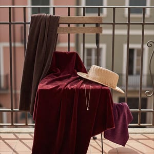 May include: A wooden chair draped with a deep burgundy velvet fabric, a dark brown velvet cloth, and a smaller purple velvet piece. A straw boater hat rests on the fabric, accompanied by a gold chain necklace with a pendant and several gold bangles.