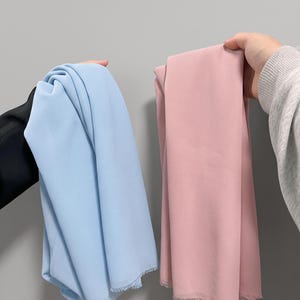 May include: Two rectangular pieces of fabric held up, one in light blue and one in dusty pink. Both pieces of fabric feature a smooth, matte texture and have frayed, unfinished edges along the bottom hem, showing a simple, lightweight textile.