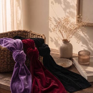 May include: Three long velvet headbands in purple, deep red, and black, each featuring a central knotted detail. The headbands are draped over the edge of a woven basket, showcasing the soft, light-reflecting texture of the velvet fabric.