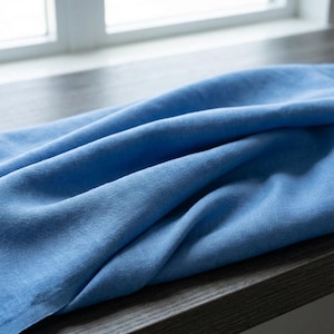 May include: A close-up shot of a folded, light blue linen fabric. The fabric is draped over a dark wooden surface, with a window visible in the background. The linen has a soft, textured appearance.