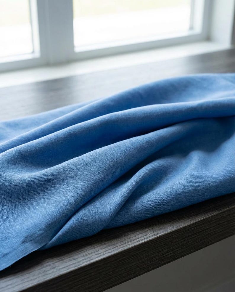 May include: A close-up shot of a folded, light blue linen fabric. The fabric is draped over a dark wooden surface, with a window visible in the background. The linen has a soft, textured appearance.