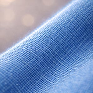 May include: Close-up of a textured, light blue fabric. The weave is visible, creating a grid-like pattern. The fabric appears soft and slightly wrinkled, with a blurred background.