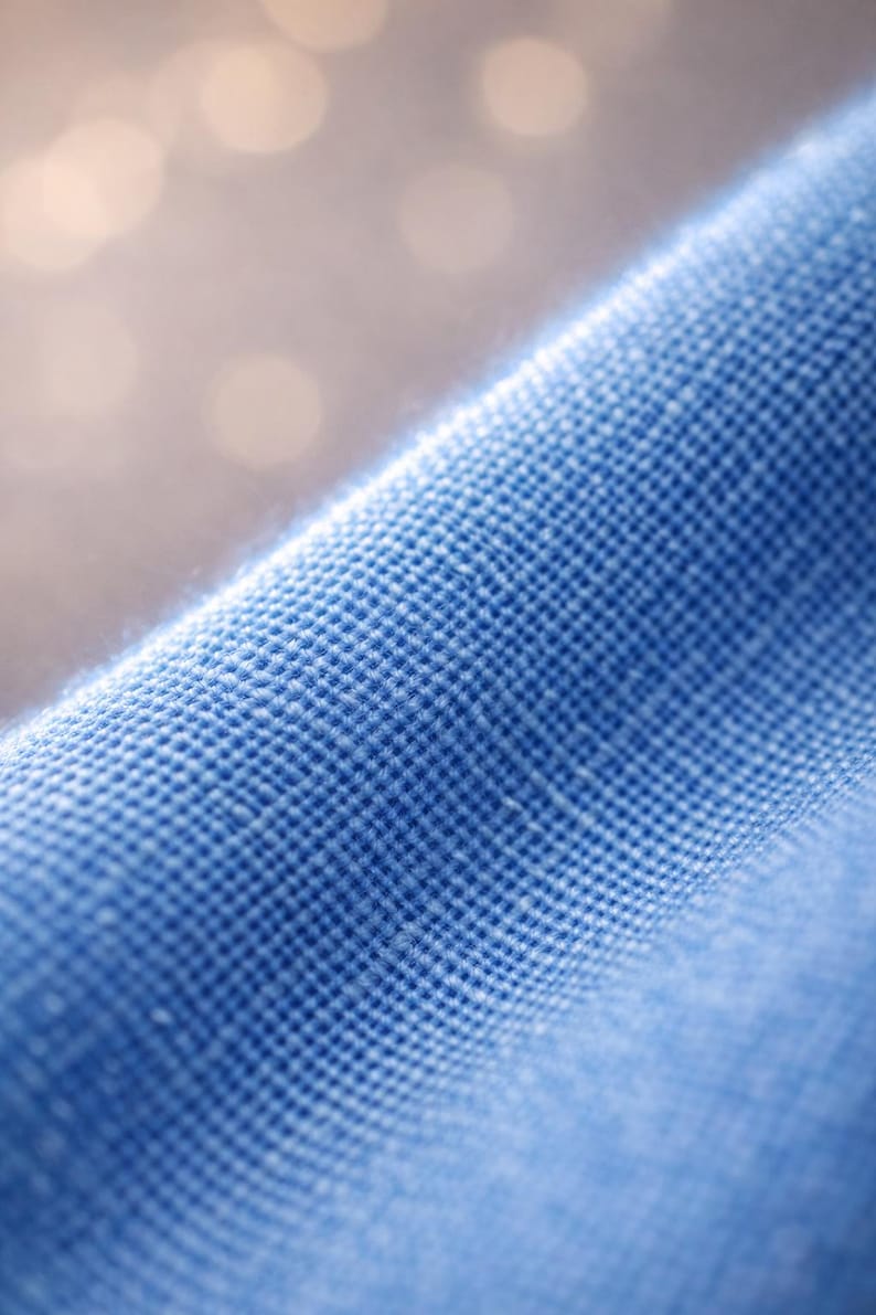 May include: Close-up of a textured, light blue fabric. The weave is visible, creating a grid-like pattern. The fabric appears soft and slightly wrinkled, with a blurred background.
