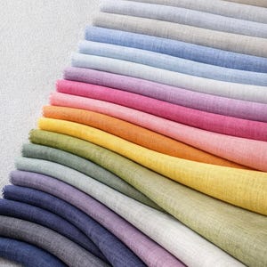 May include: A collection of folded fabric swatches in a spectrum of colors, including shades of blue, pink, orange, yellow, and green. The fabric appears to be linen or a similar material, with a slightly textured surface. The swatches are arranged in a diagonal pattern.
