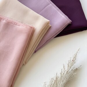 May include: A collection of four folded fabric pieces in varying shades of pink, light beige, lavender, and deep plum. Each piece of fabric features a raw, frayed edge along the top, showcasing a soft, lightweight texture and solid colour finish.