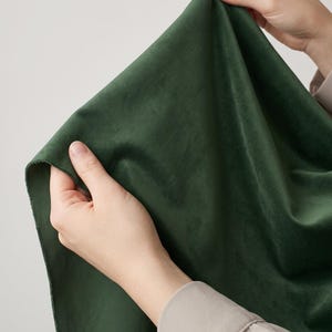 May include: A piece of dark forest green fabric with a soft, short-pile velvet texture. The material is held up to display its drape and smooth, matte surface, showing a consistent deep green color throughout the entire visible textile piece.
