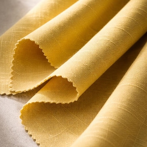 May include: Close-up of folded yellow fabric with a textured weave. The fabric has a scalloped edge and is arranged in soft folds, creating shadows and highlights. The color is a warm, golden yellow.