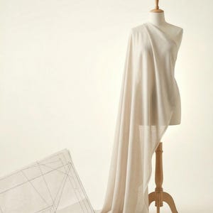 May include: A beige fabric draped over a dress form, with a pattern sheet in the foreground. The dress form is on a wooden stand. The fabric cascades down, creating soft folds and shadows.