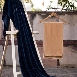 May include: A long, deep navy blue velvet fabric drape hangs over a light-colored wooden easel, pooling into a woven basket at the base. Beside it, a rectangular piece of tan-colored velvet fabric hangs neatly from a wooden clothes hanger.