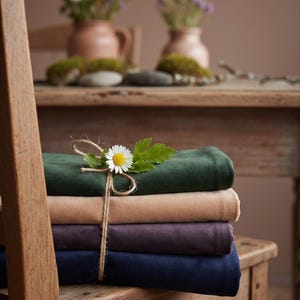 May include: A stack of four folded fabric cloths in dark green, beige, dark purple, and navy blue. The stack is tied with a thin jute twine string and decorated with a single white daisy flower and a small green leaf resting on the top cloth.