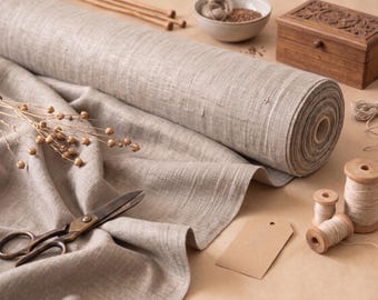 May include: A large roll of light brown, textured fabric with a visible weave, accompanied by metal scissors, three wooden spools of thread, a blank rectangular paper tag, a small wooden box, and a bowl containing small seeds.