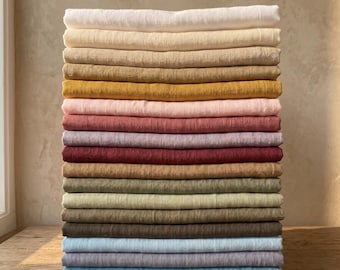 May include: A pile of folded linen textiles in a range of colours, from cream and beige to mustard, pink, and various shades of green and blue. The fabrics are stacked on a wooden surface, with lavender and a pebble in the foreground.
