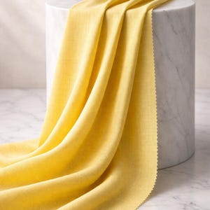 May include: A draped piece of bright yellow fabric with a textured weave. The fabric is draped over a white marble cylinder, showcasing its flowing folds and a scalloped edge. The fabric's color is a vibrant, sunny yellow.