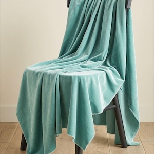 May include: A soft, light teal velvet throw blanket draped over a black wooden chair. The fabric features a smooth, matte texture with visible folds and raw, unfinished edges that hang down from the seat and backrest of the chair.